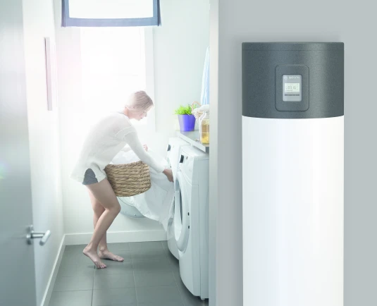 Eine Warmwasser-Wärmepumpe steht in einem Haushaltsraum. Im Hintergrund räumt eine Frau eine Waschmaschine ein.