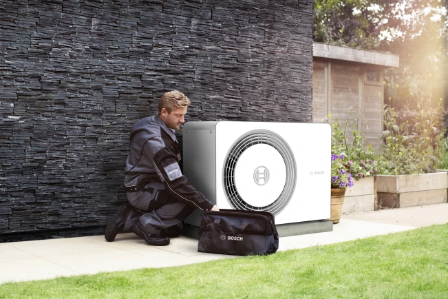 Installateur kniet neben einer Wärmepumpe von Bosch, die an einer Hauswand steht.