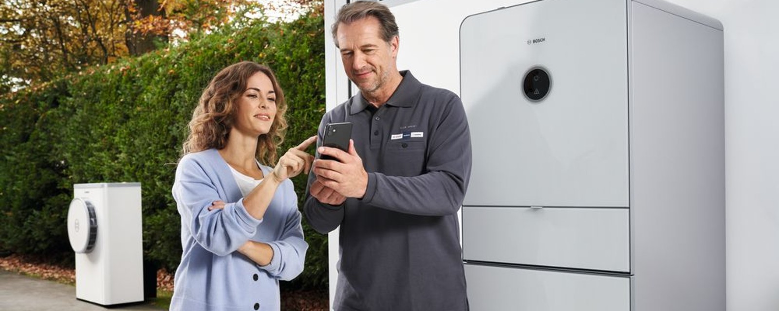Eine Frau und ein Mann stehen vor der Inneneinheit einer Wärmepumpe Compress 7400i AW und im Hintergrund sieht man die Außeneinheit stehend.. Eine Frau und ein Mann stehen vor der Inneneinheit einer Wärmepumpe Compress 7400i AW und im Hintergrund sieht man die Außeneinheit stehend..