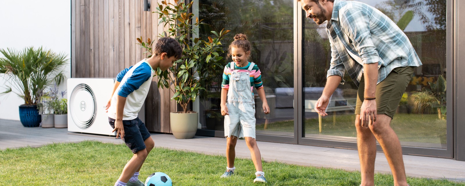 Mann und zwei Kinder spielen im Garten Fußball, im Hintergrund ist eine Wärmepumpe von Bosch an der Hauswand zu sehen. Mann und zwei Kinder spielen im Garten Fußball, im Hintergrund ist eine Wärmepumpe von Bosch an der Hauswand zu sehen.