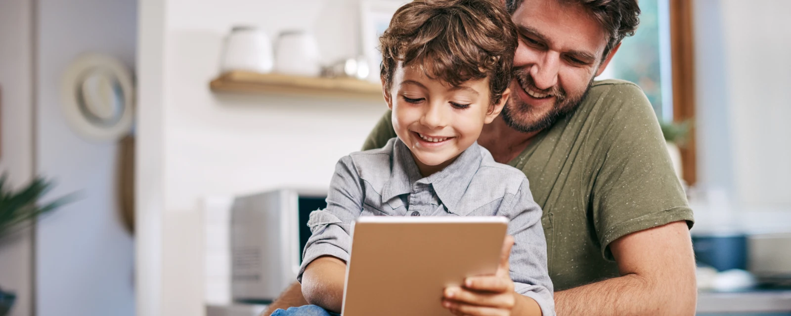 Ein Mann und ein Kind sitzen gemeinsam in einer Küche und schauen auf ein Tablet.