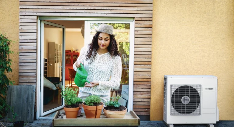 Eine Wärmepumpe Compress 3400i steht an einer Hauswand und im Vordergrund gießt eine Frau Pflanzen.