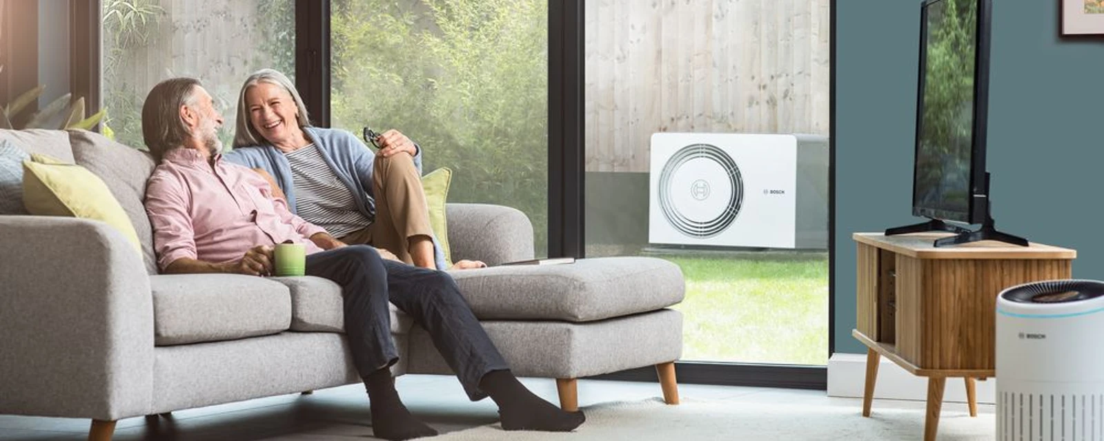 Eine Frau und ein Mann sitzen auf dem Sofa im Wohnzimmer, draußen im Garten steht eine Wärmepumpe
