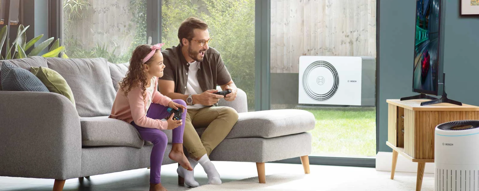 Vater und Tochter spielen gemeinsam ein Videospiel im Wohnbereich, in welchem auch ein Luftreiniger von Bosch zu finden ist, im Hintergrund auf der Terasse steht die Bosch Luft-Wasser-Wärmepumpe Compress 6800i AW.
