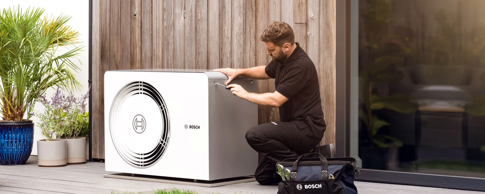 Ein Heizungsinstallateur kniet vor einer Bosch Luft-Wasser-Wärmepumpe Compress 6800i AW Außeneinheit vor einer Hauswand