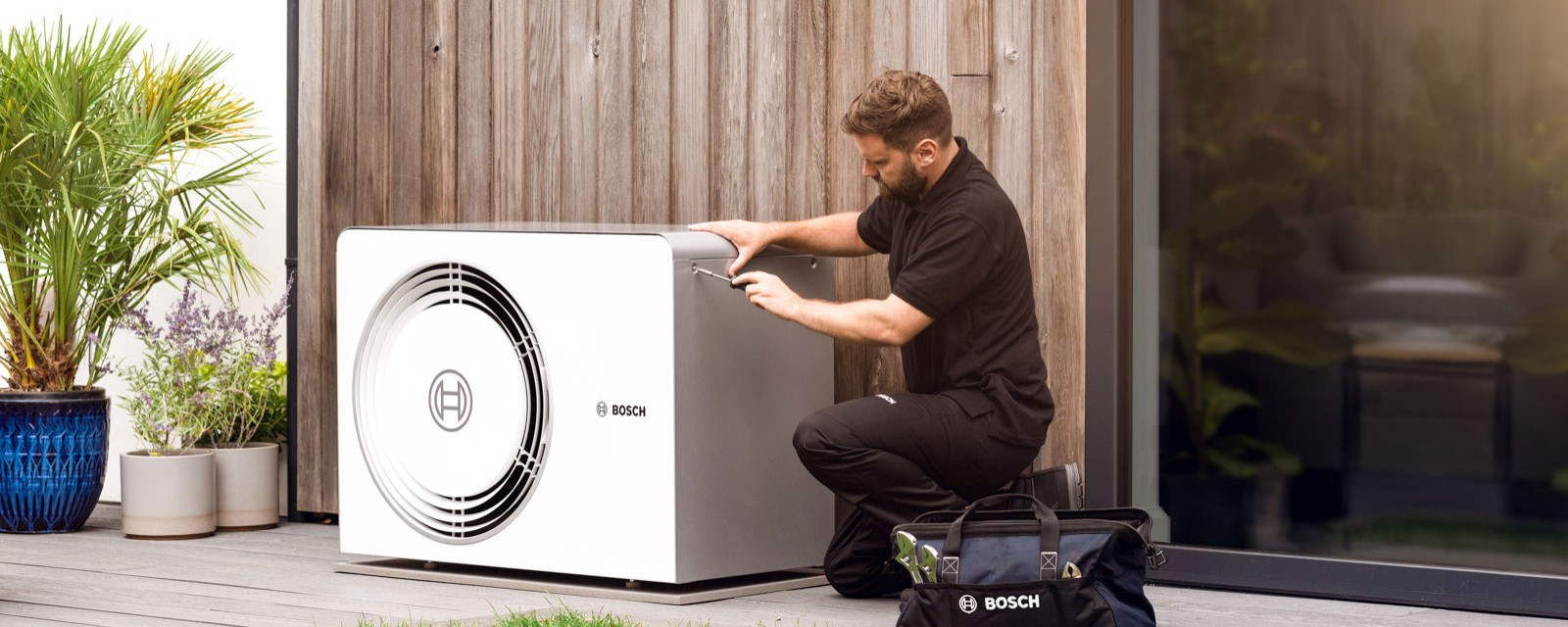 Ein Heizungsinstallateur kniet vor einer Bosch Luft-Wasser-Wärmepumpe Compress 6800i AW Außeneinheit vor einer Hauswand Ein Heizungsinstallateur kniet vor einer Bosch Luft-Wasser-Wärmepumpe Compress 6800i AW Außeneinheit vor einer Hauswand