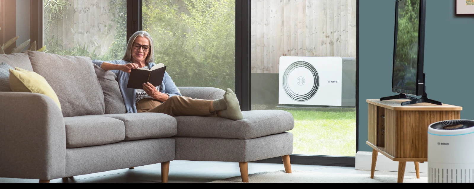 Eine Frau sitzt lesend auf einem Sofa und im Hintergrund ist eine Wärmepumpe von Bosch zu erkennen.
