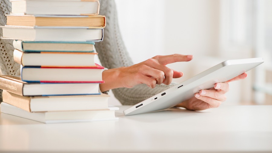 Eine Frau bedient ein Tablet, daneben steht ein Bücherstapel. Eine Frau bedient ein Tablet, daneben steht ein Bücherstapel.