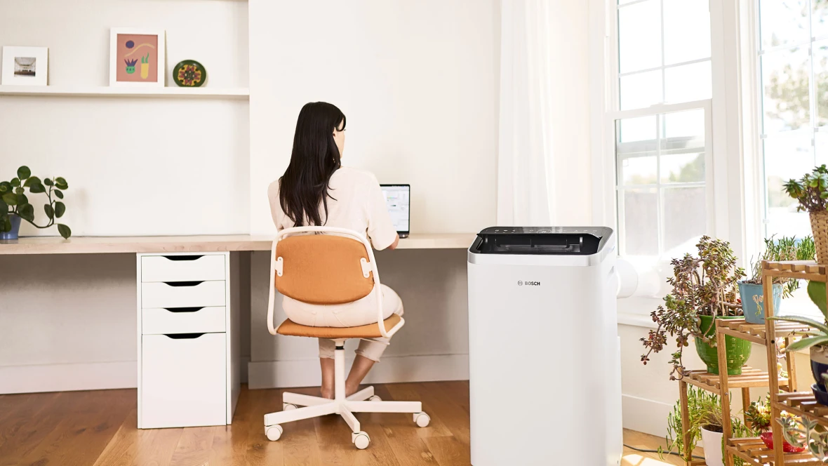 Eine Frau sitzt am Schreibtisch, während neben ihr das mobile Klimagerät Cool 5000 von Bosch steht.