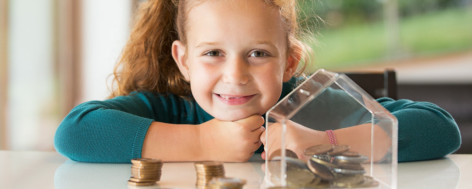 A girl smiles into the camera sitting in front of a filled money box.