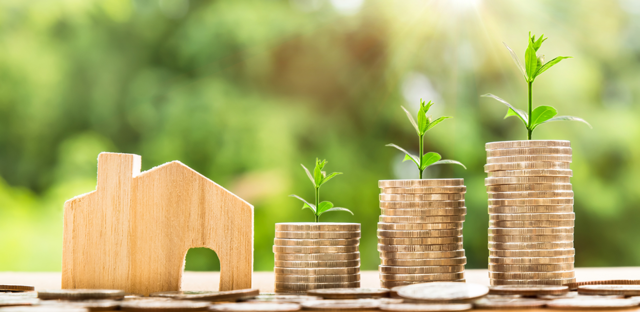A small wooden house figurine, with three piles of coins next to it. On top of the coin piles there are small green plants. A small wooden house figurine, with three piles of coins next to it. On top of the coin piles there are small green plants.