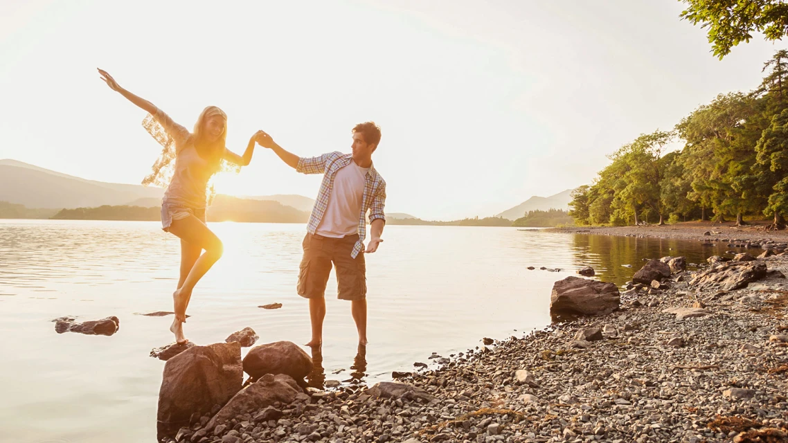 A couple walks hand in hand along a lakeshore at sunset.
