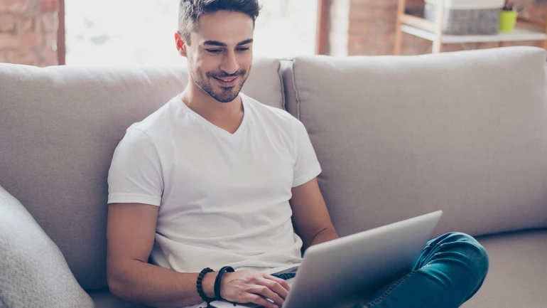 A man is sitting on the sofa with a laptop on his lap.