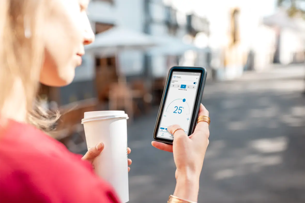 Frau steuert Bosch Compress 5800i AW Wärmepumpe per App auf dem Smartphone, hält dabei einen Kaffeebecher.