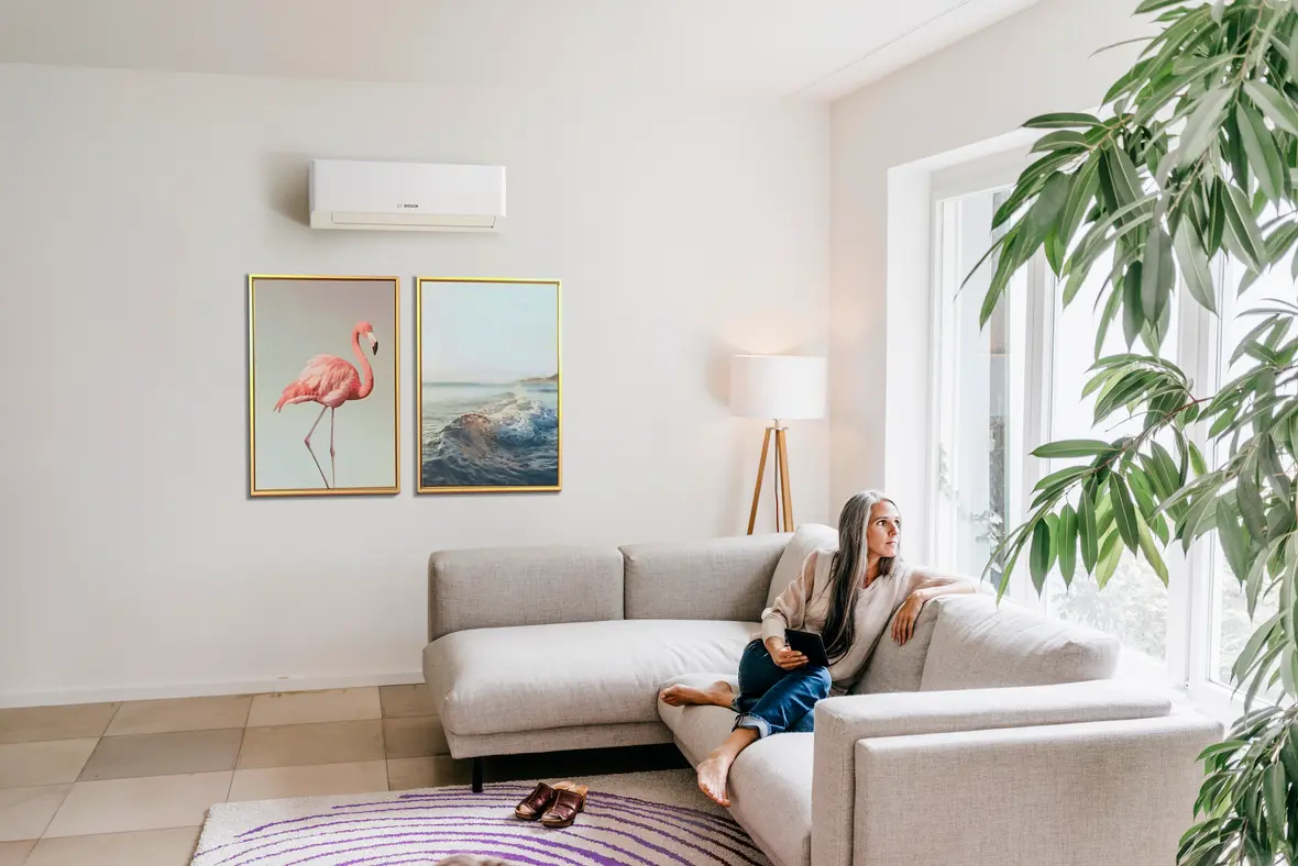 Frau auf Sofa in hellem Wohnzimmer mit Bosch Climate 7000i Single-Split Klimagerät an der Wand über zwei Bildern.