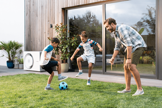 Familie spielt im Garten vor Hauswand mit installierter Bosch Compress 5800i AW Wärmepumpe.
