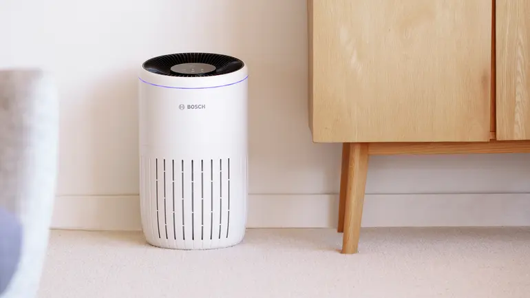 Der Bosch Air 4000 Luftreiniger in einem Wohnambiente. Das Gerät steht auf einem hellen Teppichboden neben einem Holzschrank.