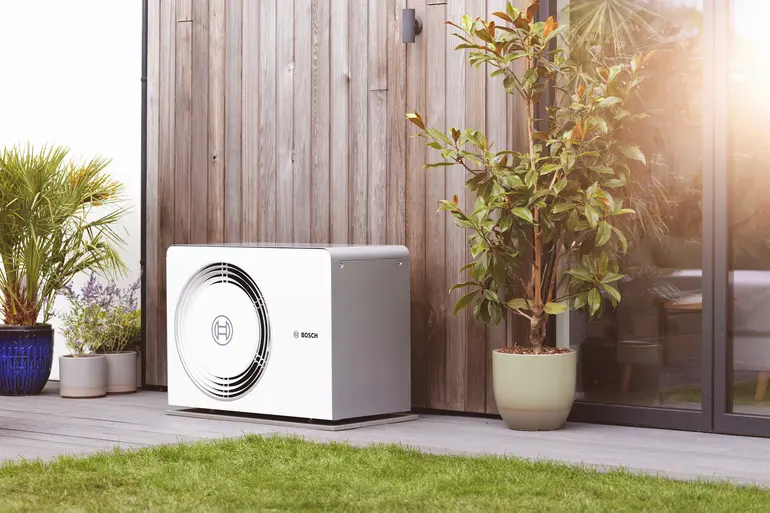 Bosch Compress 5800i AW Wärmepumpen Außeneinheit auf Terrasse vor Holzfassade, umgeben von Pflanzen und Glasfront bei Sonnenschein.