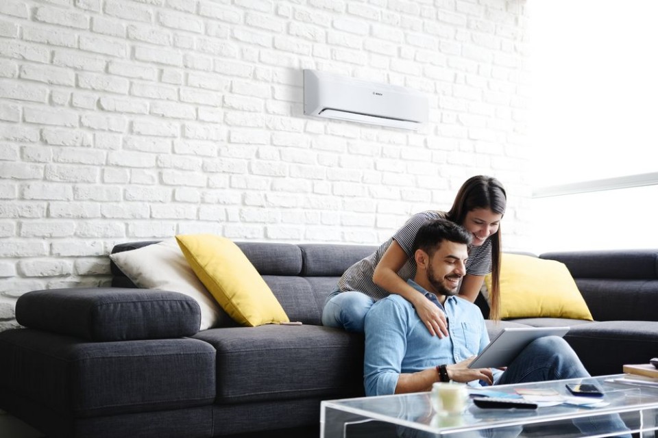 The picture shows a man and a woman sitting together on a cosy sofa. A modern air conditioning unit is mounted on the wall in the background.
