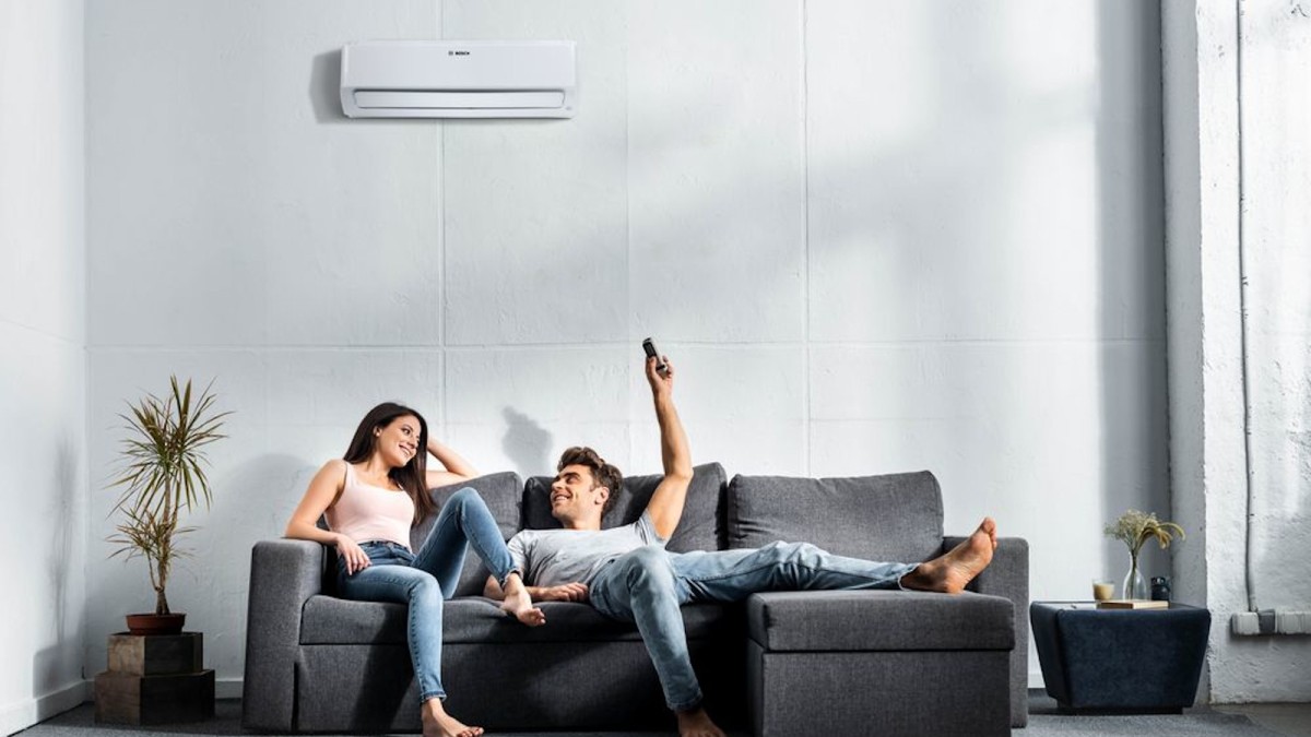 Couple sitting comfortably on the sofa, man operates the Bosch Climate Class 6000i on the wall with the remote control.