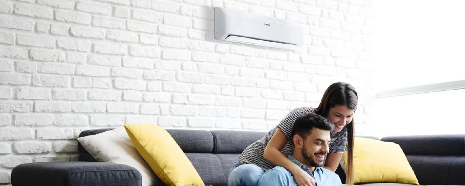 The picture shows a man and a woman sitting together on a cosy sofa. A modern air conditioning unit is mounted on the wall in the background.