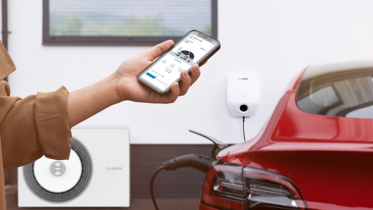 Ruka držiaca mobil s otvorenoi aplikáciou Bosch HomeCom Easy; nabíjajúce sa auto, wallbox Bosch a tepelné čerpadlo Bosch v pozadí Ruka držiaca mobil s otvorenoi aplikáciou Bosch HomeCom Easy; nabíjajúce sa auto, wallbox Bosch a tepelné čerpadlo Bosch v pozadí