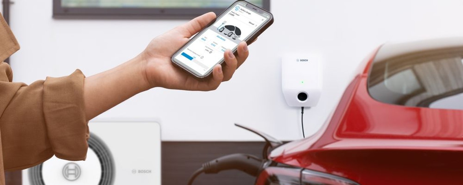 Detail na ruku držiacu mobil s otvorenou aplkáciou pre nabíjanie elektrického auta, wallbox Bosch nabíjajúci červené auto v pozadí