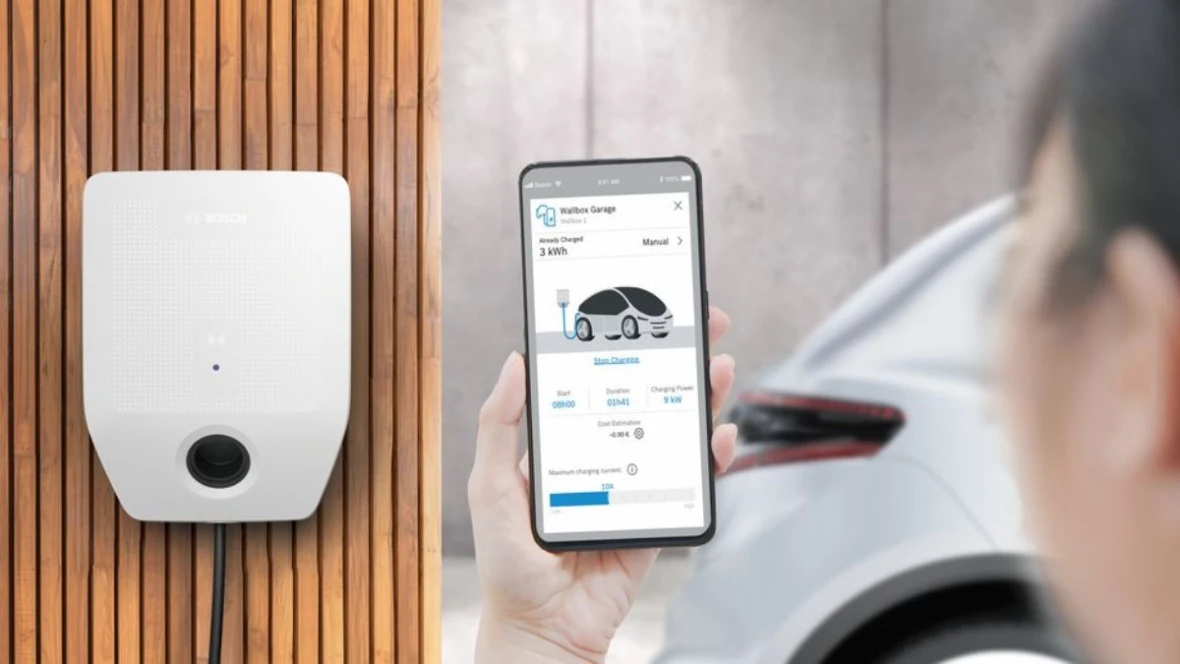 Detail na obrazovku mobilného telefónu v ruke použítaveľa stojaceho pre wallboxom Bosch, aplikácie zobrazuje stav nabitia elektromobilu, zadná časť bieleho elektromobilu v pozadí
